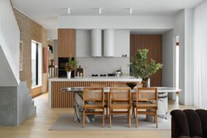 An open concept space in a modern cottage, looking past the marble dining table and rattan dining chairs into the kitchen. Featuring lots of fresh greenery, timber panelling on the kitchen island, and white tiling on the backsplash and overhead.