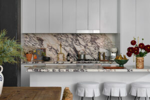 A European-inspired eclectic warehouse apartment kitchen with vintage timber furniture, wrought iron upholstered stools, vintage brass finishings, and Italian marble backsplash and bench tops.