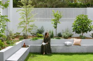 The exterior of a modern Queenslander, the homeowner sits on large concrete steps surrounded by lush greenery and landscaping.