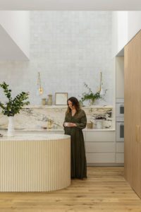 A modern kitchen with a rounded kitchen island with fluted edging, a marble backsplash, and styled open shelving. The homeowner stands at the bench holding a mug.