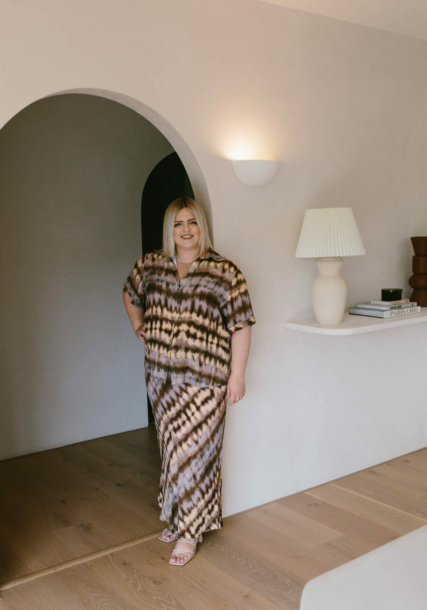 Hayley Jenkin stands in an archway with a lamp and objects on a shelf to her right.